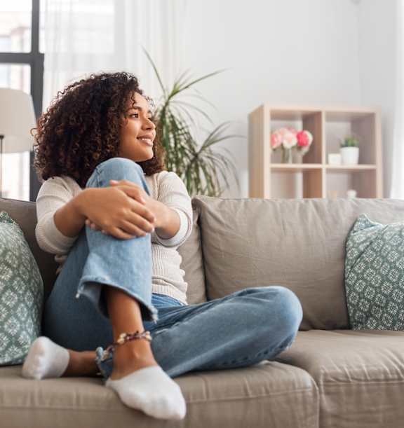 A woman with curly hair is sitting on a beige couch with a blue pillow, wearing a white top and blue jeans, with her legs crossed and holding a blue object.