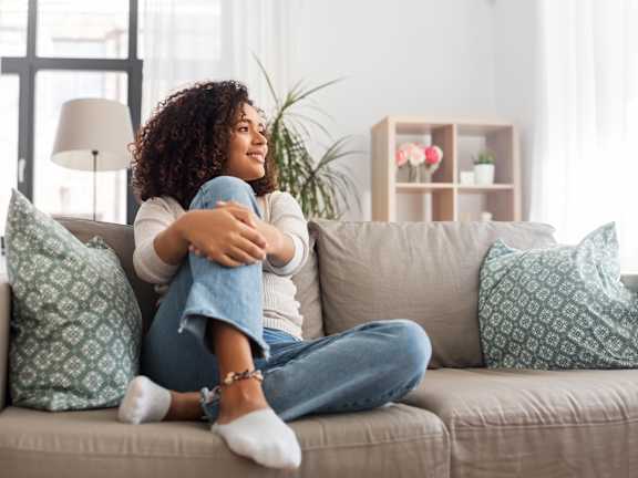 A woman with curly hair is sitting on a beige couch with a blue pillow, wearing a white top and blue jeans, with her legs crossed and holding a blue object.