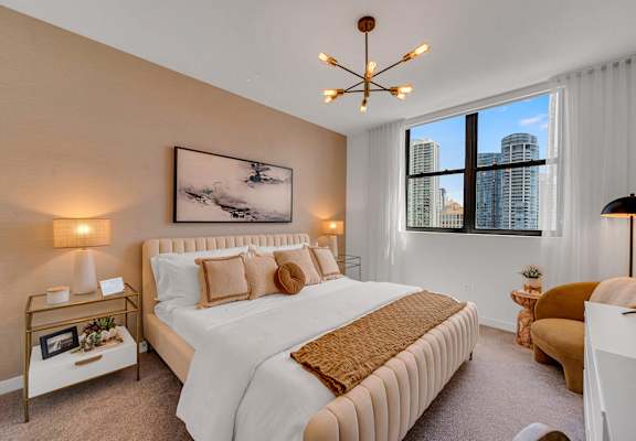 A bedroom with a large bed, a painting on the wall, and a window with a view of the city.