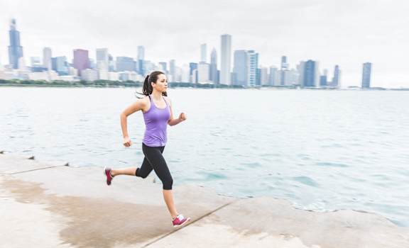 A woman in a purple top and black leggings runs along a concrete path by the water.