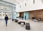 Thumbnail 11 of 12 - A man is walking through a spacious lobby with people standing in the background.