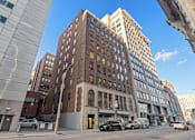 Thumbnail 7 of 7 - A tall brown building with many windows is in the middle of a city street.