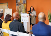 Thumbnail 2 of 16 - A woman is giving a speech at a podium in front of a group of people.