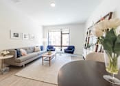Thumbnail 12 of 82 - Apartments for Rent in Beverly, MA - Holmes Beverly - Expansive Living Room with Bright Lighting, Grey Sofa, Blue Armchairs, Wooden Coffee Table, White Rug Over Wood Flooring, Framed Art, White Side Tables, Standing Lamp, White Walls, White Storage Monitor, Wood and White Storage Shelving Rack, and a Large Window