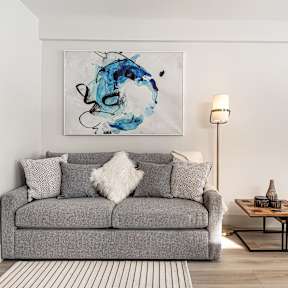 A grey couch with a white pillow and a blue painting on the wall.