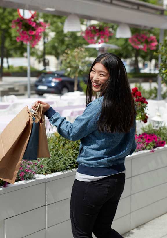 A woman is holding a brown paper bag and smiling while standing on a sidewalk.