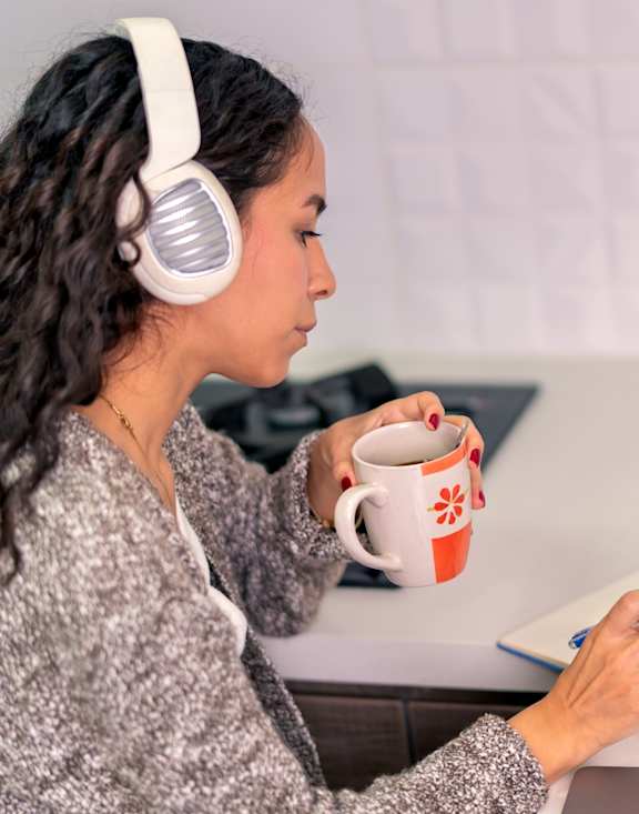 A woman with headphones on is reading a book while holding a mug.