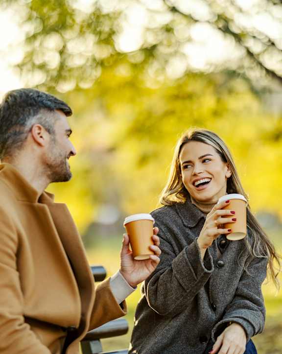 A man and a woman are sitting on a bench in a park, each holding a cup.
