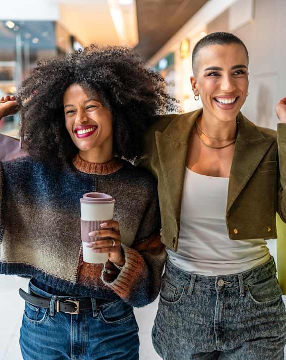 Two women are standing together, one holding a coffee cup and the other holding a yellow shopping bag.