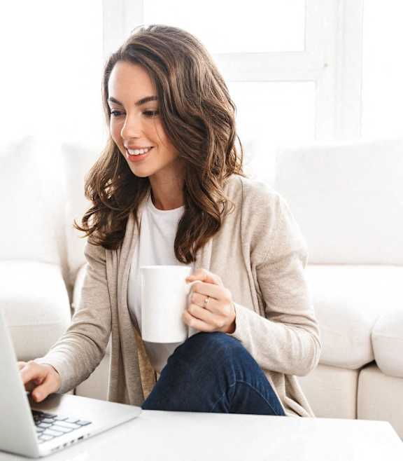 A woman is sitting on a couch with a laptop and a mug in her hand.