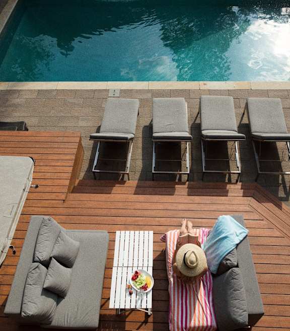 A person is sitting on a deck by a pool with a hat on.