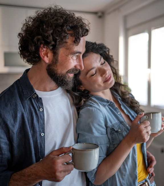 A man and a woman are standing close together, each holding a mug, in a room with a window.