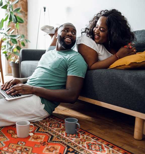 A man and woman are sitting on the floor in front of a couch, with the man using a laptop and the woman resting her head on his shoulder.