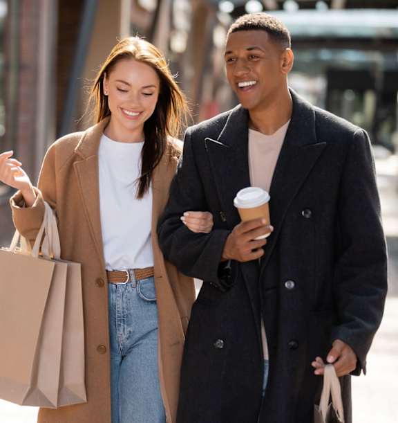 A man and a woman are walking down the street, the woman is holding a brown paper bag and the man is holding a coffee cup.