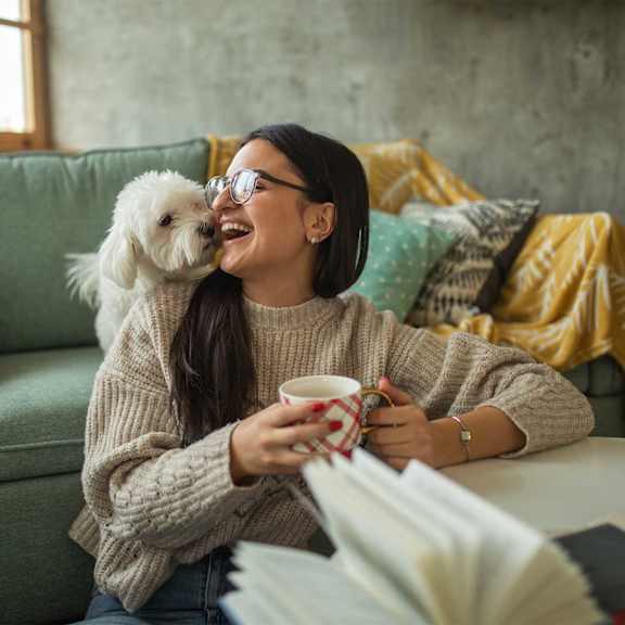 A woman is sitting on a couch with a dog, holding a cup, and reading a newspaper.