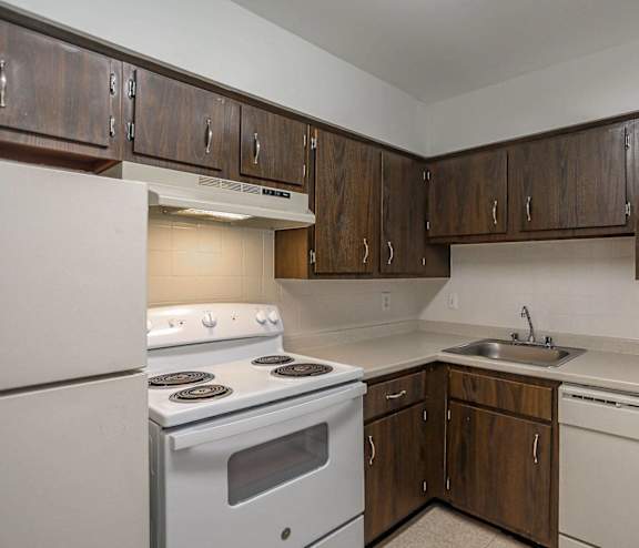 A kitchen with white appliances and wooden cabinets, disposals in each apartment, electric ranges, and dishwashers.