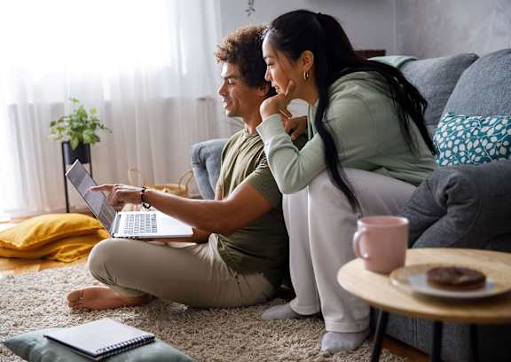 A man and woman are sitting on the floor in front of a couch, with the man using a laptop and the woman holding a cup.