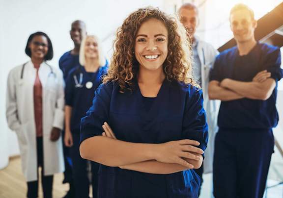 A group of medical professionals, including a woman in a blue scrub, stand in a line with their arms crossed.