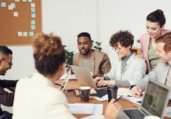 A group of people are gathered around a table with laptops, engaged in a discussion or meeting.