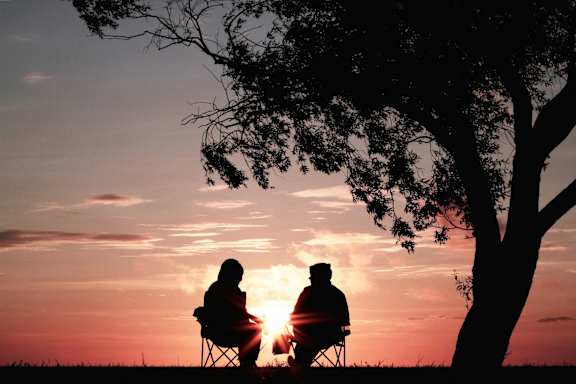 two people sitting under a tree watching the sunset