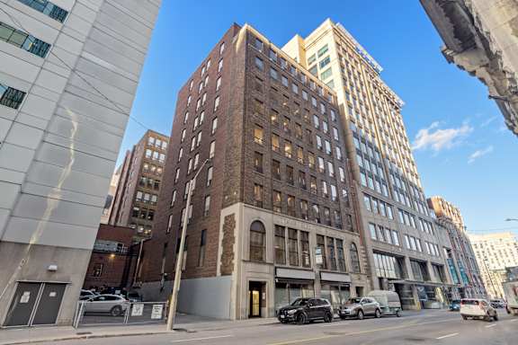 A tall brown building with many windows is in the middle of a city street.