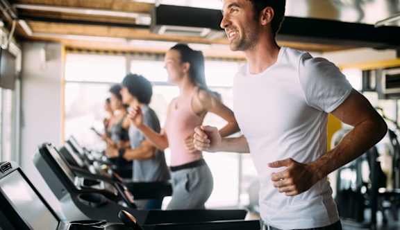 A man in a white t-shirt is running on a treadmill in a gym with other people exercising around him at The Legacy Apartments in Seminole Heights, FL