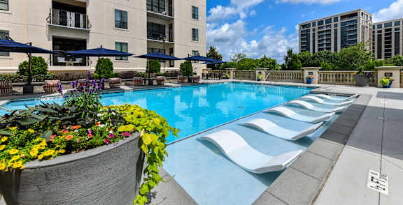 A large swimming pool with a concrete edge and a concrete planter with colorful flowers in front of apartment buildings.