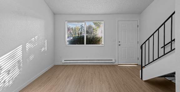 A room with a wooden floor, a window with blinds, a white door, and a staircase with a black railing.