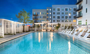 a swimming pool with white chaise lounge chairs and an apartment building in the background
