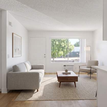 A living room with a grey couch, a coffee table, and a window with a view of trees and buildings outside.