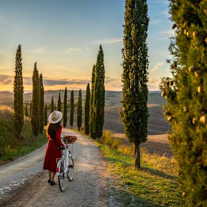 A woman in a red dress and straw hat rides a bicycle down a dirt road lined with tall green cypress trees.