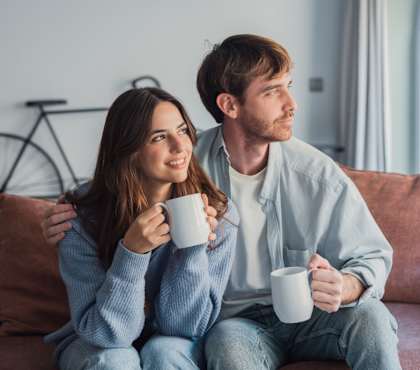 A man and a woman are sitting on a couch, each holding a white mug.