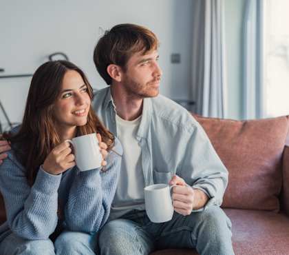 A man and a woman are sitting on a couch, each holding a white mug.