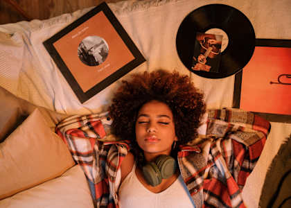 A woman with curly hair is lying on a bed with a plaid shirt and headphones around her neck.