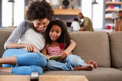 Woman and girl reading a book while smiling, man with boy at computer in background.