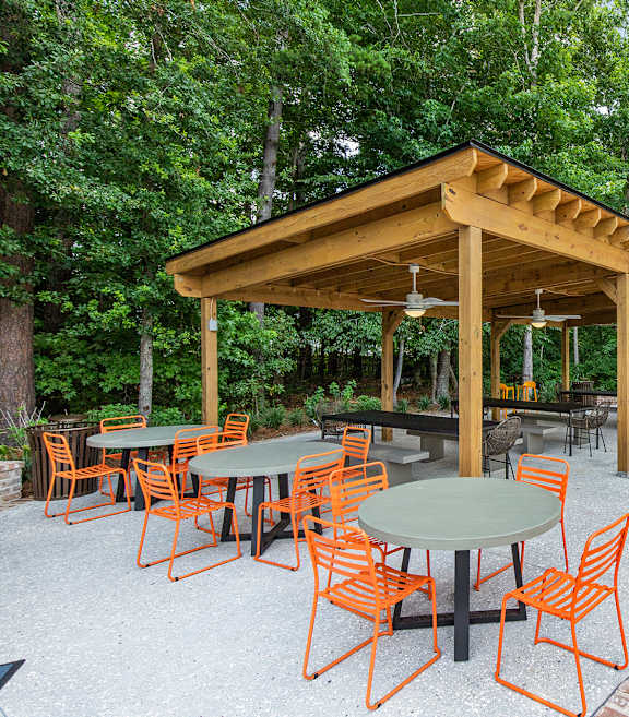 A wooden pavilion with orange chairs and a green table is surrounded by trees.