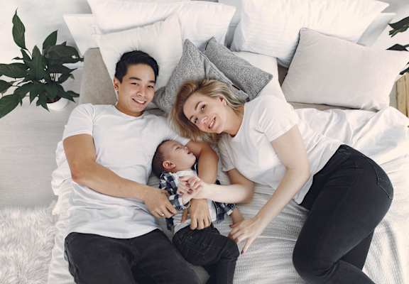 A family of three is lying on a bed with pillows and a plant in the background.