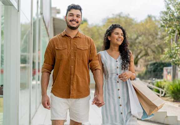 A man and a woman are walking together, the man is wearing a brown shirt and white shorts, and the woman is carrying a brown paper bag.
