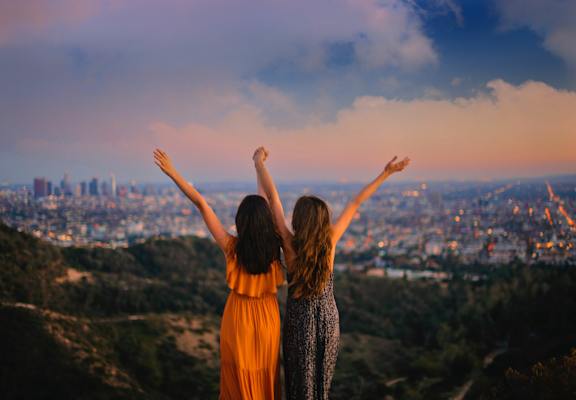 Two women with arms raised in a cityscape at sunset.