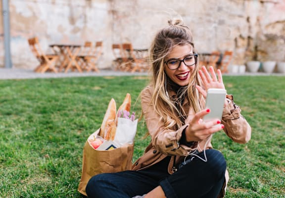 A woman is sitting on the grass with a bag of food and a phone in her hand.