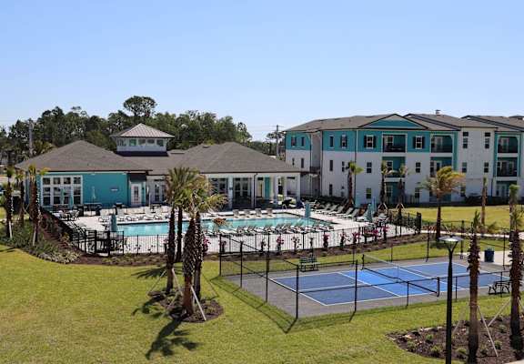 A tennis court is surrounded by a pool and palm trees.