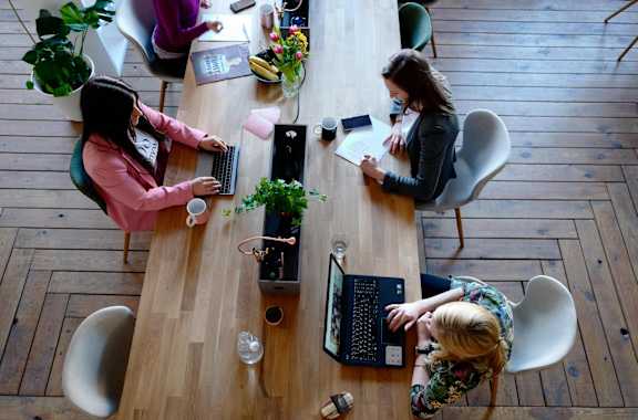 Three women are working at a wooden table with laptops and notebooks.