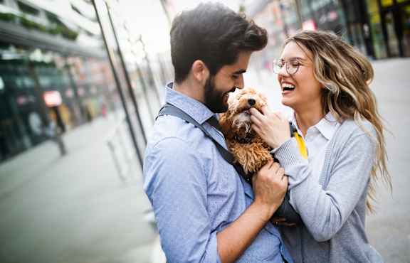 Man and Woman Standing on Sidewalk and Smiling while Holding Dog