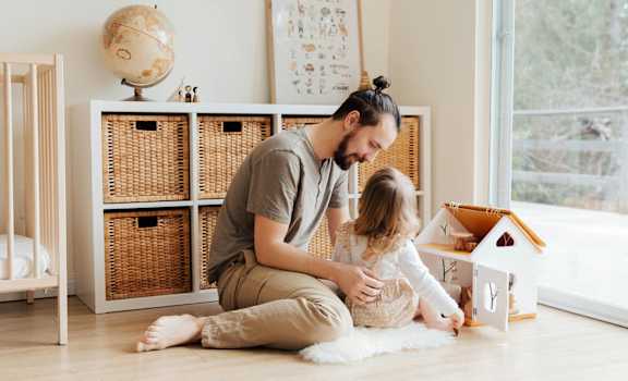 A man and a little girl are playing with a wooden dollhouse on the floor.