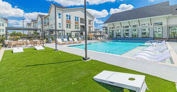 A white lawn chair is on a green lawn in front of a pool and a building.