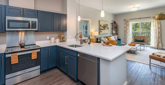 The Residences at Crosspoint open-plan kitchen and living area includes navy blue shaker cabinets, stainless steel appliances, a white island countertop with stools, and seating near a sliding glass door leading to a balcony.