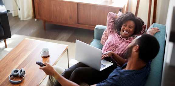 A man and woman are sitting on a couch with a laptop and a remote control in hand. at The Glen, Texas, 75067