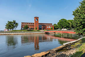 Brick waterfront museum with tower reflected in the river, connected by a red pedestrian bridge beneath a clear sky.