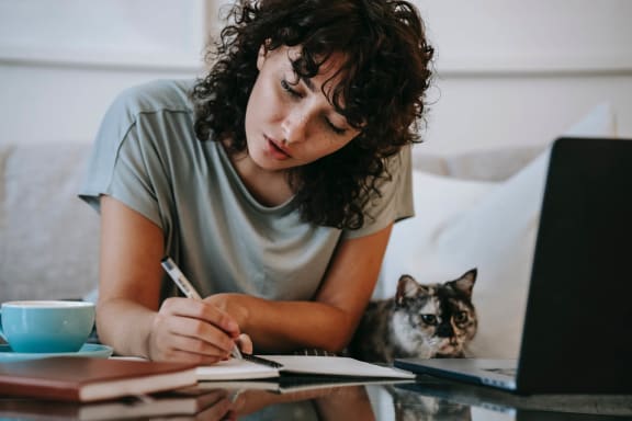 a woman sitting at a table writing in a notebook with a cat next to her at Pinehurst Condominiumse Apartments ,Las Vegas, Nevada, 89118