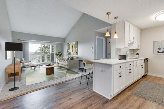 A modern kitchen with white cabinets, hanging lights, stainless steel appliances, and wood-like floors. In the back, there is a vaulted ceiling over a spacious carpeted living room with a balcony.